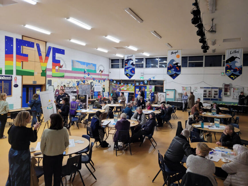 Image from the Ballymun Community Conference run by Codema. A school sports hall if full of people sitting at round tables, there are colourful posters on the wall in the background