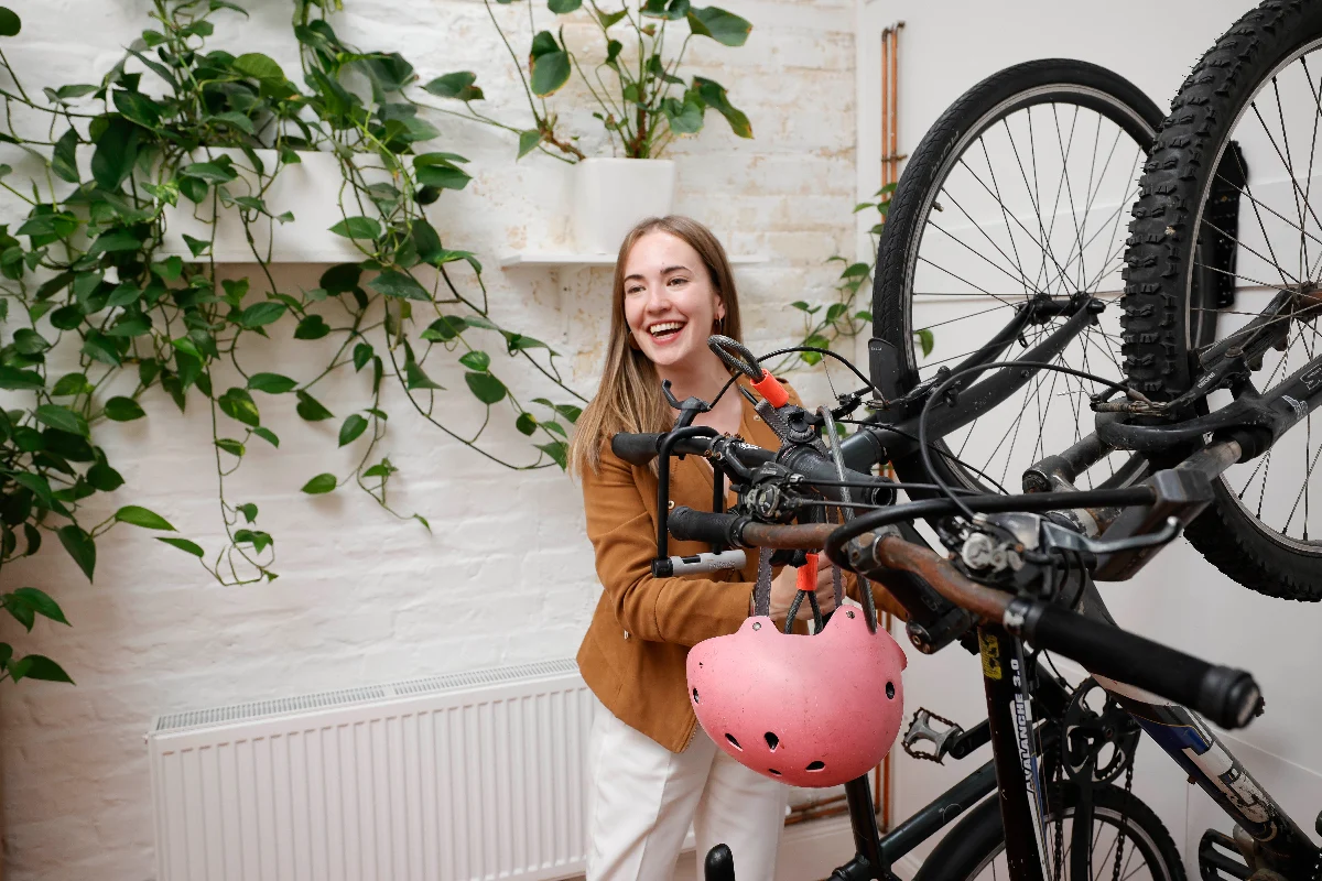 Codema staff member. A woman takes her bike down from the bike storage on the wall. There are two bikes hanging and a pink helmet. There are plants in the background.