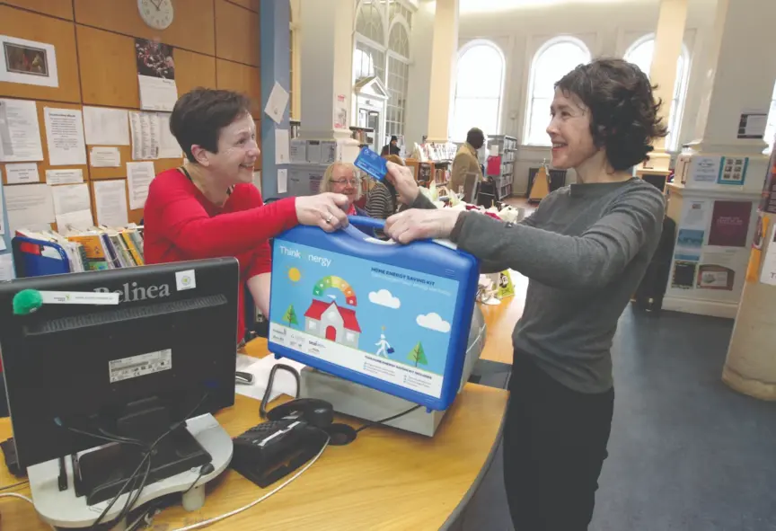 Image of two women in a library, one is handing a blue Home Energy Saving Kit over the counter to the other