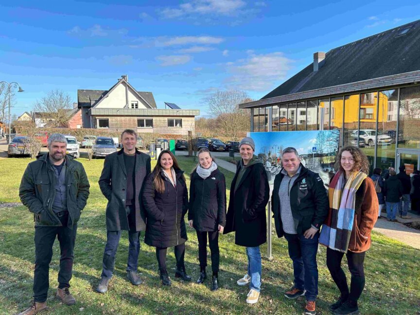 Image of a group of people standing in a row smiling at the camera. They are standing on grass with buildings and blue sky behind them.