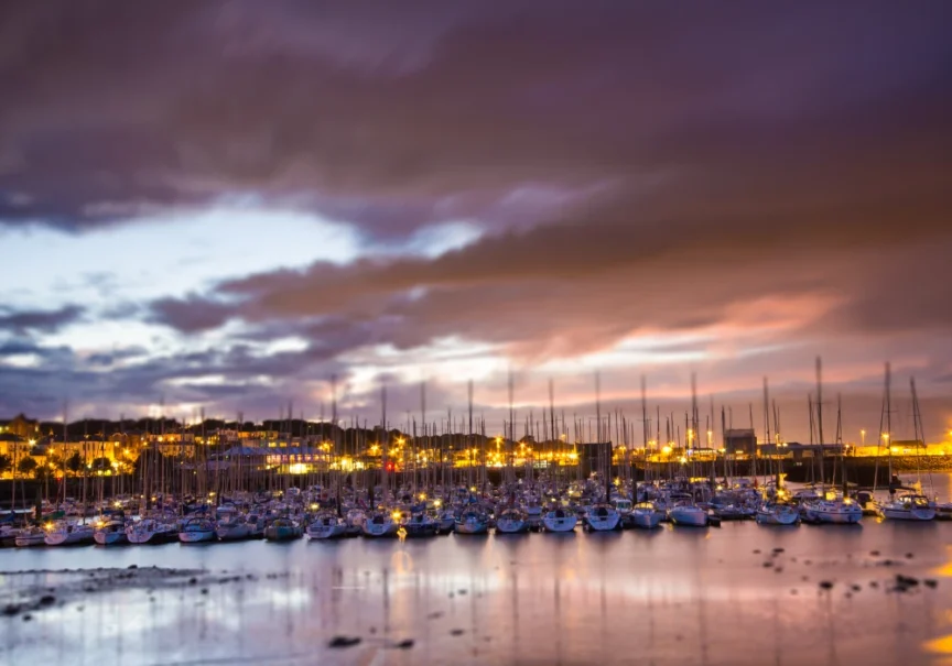 Image of boats in a harbour at dusk with lights behind them