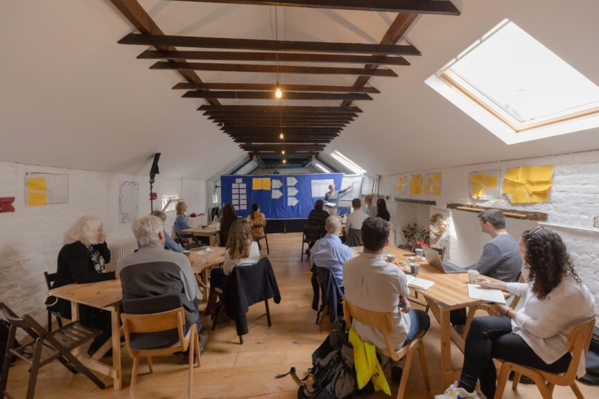 Image of a workshop taking place in a room with white walls and exposed wooden beam ceiling. Participants are sitting around tables looking at the facilitator at the top of the room